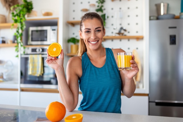 Schöne junge Frau, die frischen Orangensaft in der Küche trinkt Gesunde Ernährung Glückliche junge Frau mit Glas Saft und Orange am Tisch in der Küche