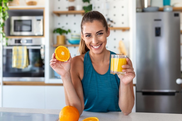 Schöne junge Frau, die frischen Orangensaft in der Küche trinkt Gesunde Ernährung Glückliche junge Frau mit Glas Saft und Orange am Tisch in der Küche