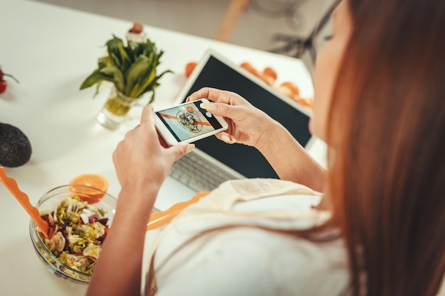 Schöne junge Frau, die gesunden Salat mit Smartphone für ihren Vlog fotografiert.