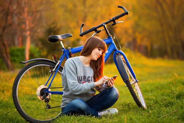 Schöne junge Frau, die Handy verwendet, das bei Sonnenuntergang neben ihrem Fahrrad sitzt