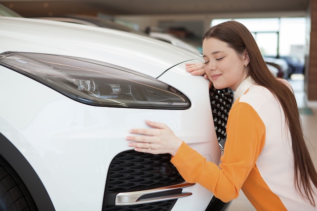 Schöne junge Frau, die ihr neues Auto im Autohaus umarmt und mit geschlossenen Augen lächelt