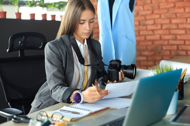 Schöne junge Frau, die ihre Notizen betrachtet und Digitalkamera beim Sitzen in ihrer Werkstatt hält.