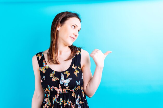 Schöne junge Frau, die ihren Daumen auf die Seite zeigt, die mit blauem Hintergrund überzeugt ist, der ein Sommerkleid trägt.