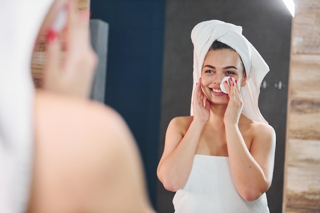 Schöne junge Frau, die im Badezimmer steht, in den Spiegel schaut und sich um ihr Gesicht kümmert.