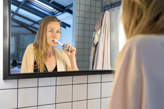 Schöne junge Frau, die in Badezimmerspiegel schaut, der ihre Zähne mit Zahnbürste putzt