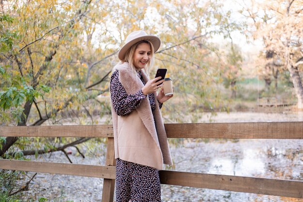 Schöne junge Frau, die Kaffee trinkt, mit Telefon in der Natur im Herbstpark im Herbst.