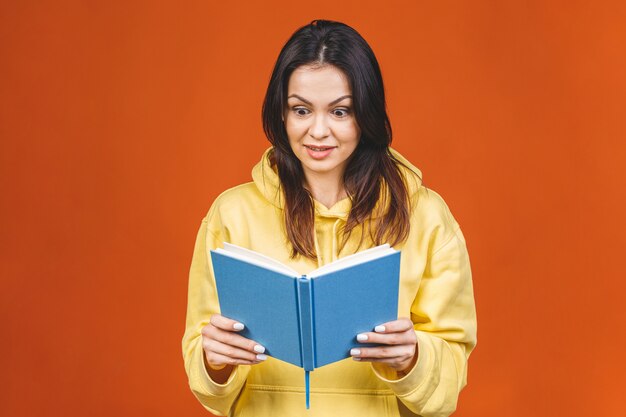 Schöne junge Frau, die lässig stehend lokalisiert über orange Hintergrund trägt und ein Buch liest.