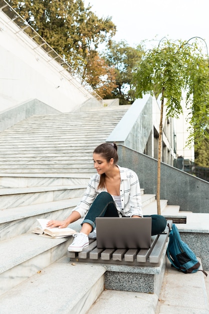 Schöne junge Frau, die Laptop-Computer im Freien beim Lesen des Buches verwendet