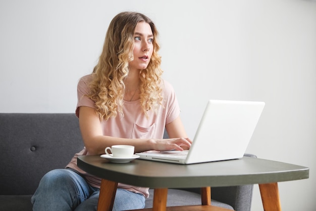 Foto schöne junge frau, die mit einer weißen tasse kaffee auf dem sofa sitzt und an einem weißen laptop arbeitet mädchen herein