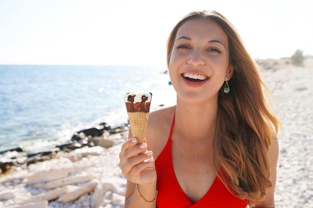 Schöne junge Frau, die mit Eistüte lächelt und am Strand in die Kamera schaut