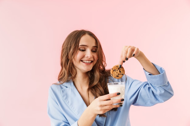 Schöne junge Frau, die Pyjamas trägt, die lokal über rosa Hintergrund stehen, Schokoladenplätzchen essen, Milch trinkend