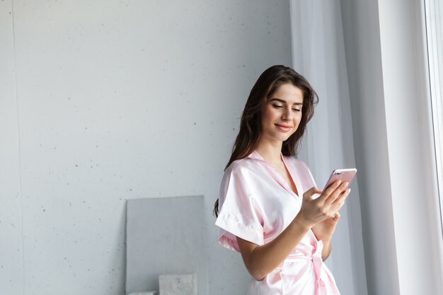 Schöne junge Frau, die Robe trägt, die am Fenster im Schlafzimmer steht und auf Handy spricht