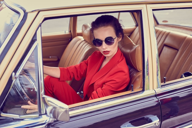 Schöne junge Frau, die rotes Kostüm und Sonnenbrille im Retro-Auto sitzt