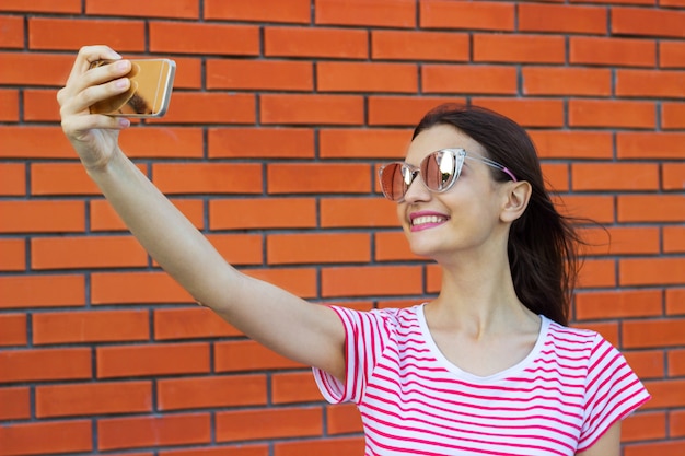 Schöne junge Frau, die Selbstporträt auf einem Smartphone auf einer Backsteinmauer macht