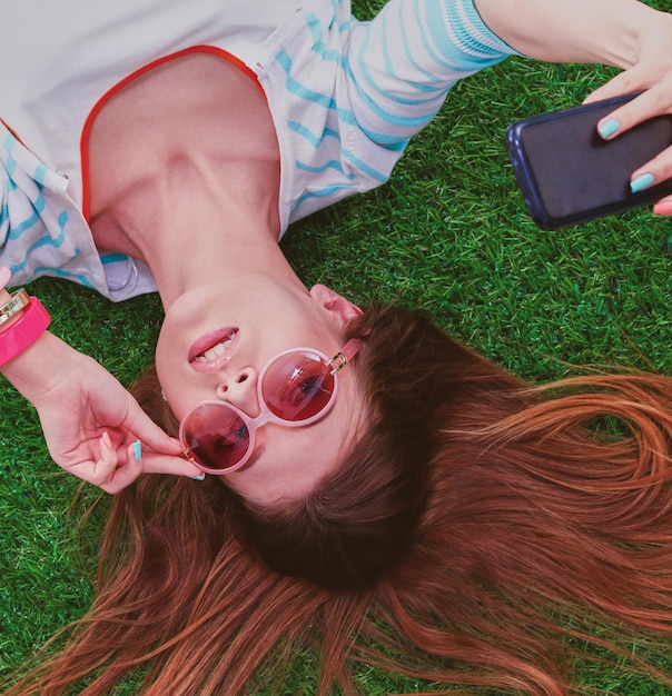 Schöne junge Frau, die Selfie mit ihrem Telefon macht, während sie im grünen Gras liegt schöne junge Frau ma