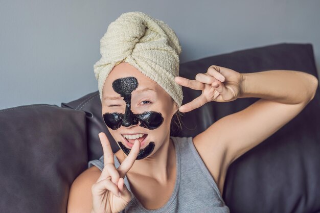 Schöne junge Frau, die sich zu Hause mit Gesichtsmaske entspannt. Glückliche fröhliche Frau, die eine schwarze Maske auf das Gesicht aufträgt