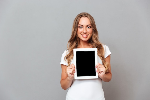 Schöne junge Frau, die Tablet-Computer mit leerem Bildschirm lokalisiert auf einer grauen Wand zeigt