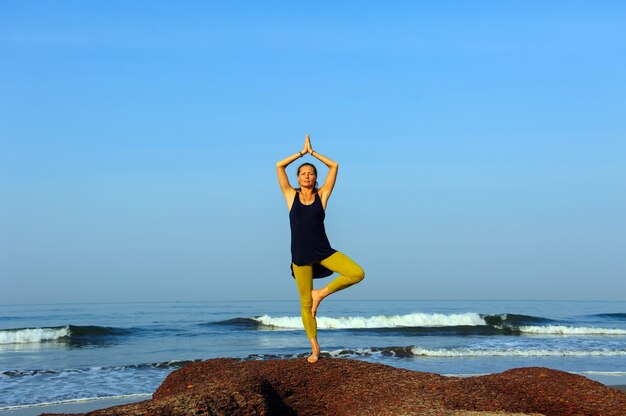 Schöne junge Frau, die Yoga und Dehnungsübungen am Sommerozeanstrand praktiziert.