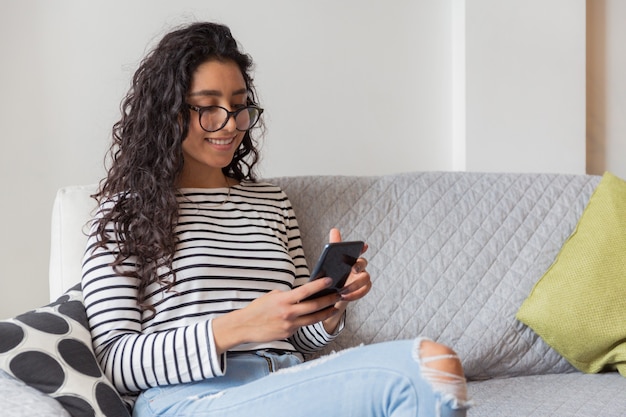 Schöne junge Frau, die zu Hause auf dem Sofa liegend genießt und auf ihrem Smartphone tippt