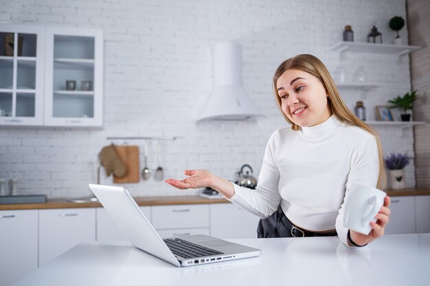 Schöne junge Frau, die zu Hause auf einem Laptop studiert. Ein Mädchen mit langen blonden Haaren in einem Pullover trinkt Tee in der Küche. Arbeiten Sie aus der Ferne von zu Hause aus