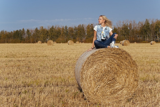 Schöne junge Frau Dorfbewohnerin posiert in Jeans auf einem Ballen Heu in einem Feld