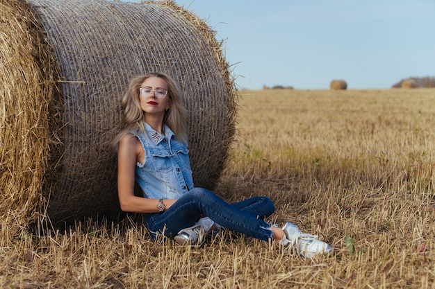 Schöne junge Frau Dorfbewohnerin posiert in Jeans nahe einem Ballen Heu in einem Feld