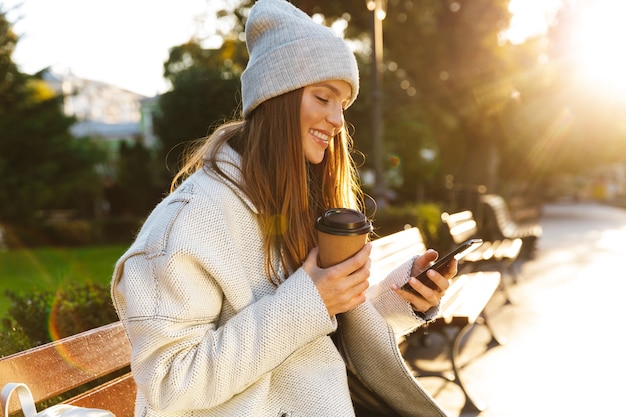 Schöne junge Frau gekleidet in Herbstmantel und Hut, die auf einer Bank draußen sitzen, Kaffee trinkend, mit Handy