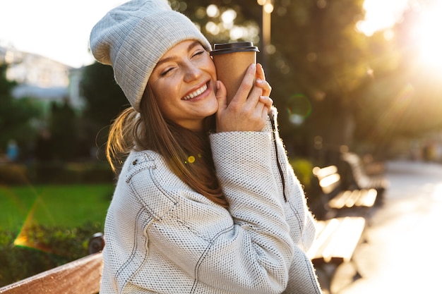 Schöne junge Frau gekleidet in Herbstmantel und Hut, die auf einer Bank draußen sitzen und Tasse warmen Kaffee halten