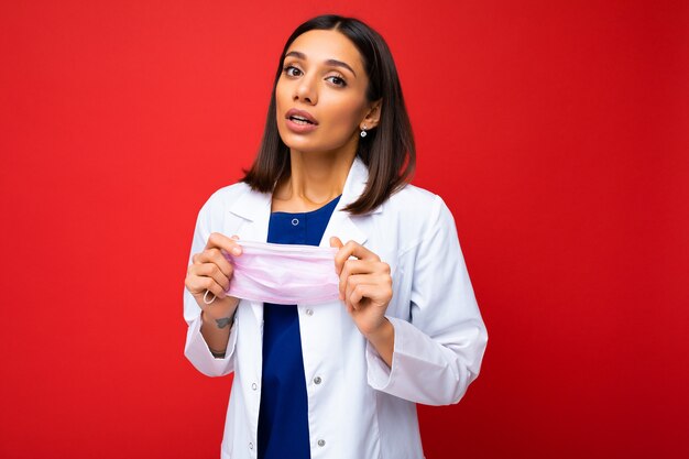 Schöne junge Frau hält und trägt eine weiße medizinische Maske, um sich vor dem Koronavirus zu schützen, kümmert sich um ihre Gesundheit und Sicherheit, hält sich an Selbstisolierung. Konzept von Covid-19