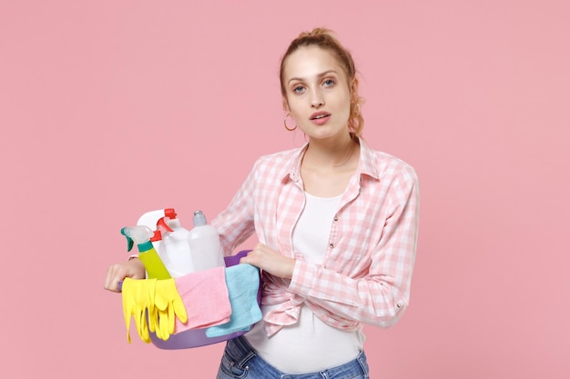Schöne junge Frau Hausfrau im karierten Hemd Haltebecken mit Waschmittelflaschen, die Reinigungsmittel waschen, während sie Hausarbeit machen, isoliert auf rosa Hintergrund Studio. Housekeeping-Konzept. Kamera suchen.