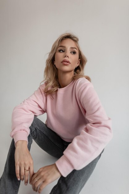 Schöne junge Frau Hipster mit blonden Haaren in einem modischen rosa Sweatshirt und grauen Vintage-Jeans sitzt im Studio auf dem Boden