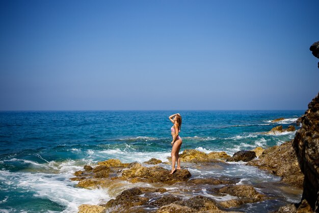 Schöne junge Frau im Badeanzug an einem felsigen Strand an einem sonnigen Tag vor dem Hintergrund der Wellen. Urlaub in der Sommersaison. Selektiver Fokus