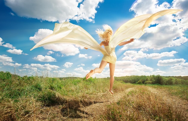 Schöne junge Frau im beigen Kleid, die auf Straßenfeld auf Hintergrund des blauen Himmels mit Kopienraum springt