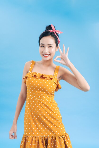 Schöne junge Frau im eleganten gelben Cocktailkleid im Tupfenmuster schaut in die Kamera, lacht und zeigt ein ok Handzeichen.