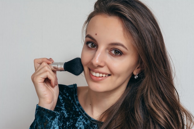 Schöne junge Frau im glänzenden grünen Kleid, das Pulver mit einem Pinsel auf das Gesicht aufträgt.