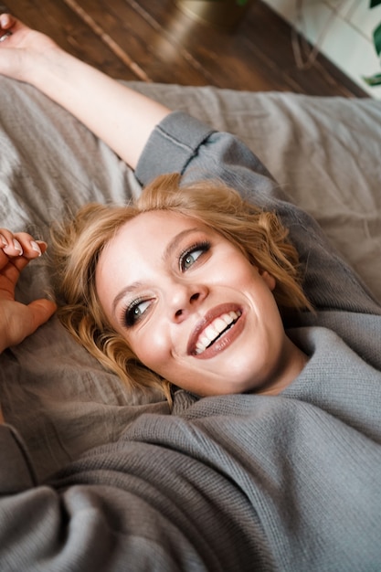 Schöne junge Frau im grauen Pullover lachend im Bett liegend in ihrem gemütlichen Schlafzimmer. Nettes Mädchen, das zu Hause entspannt. Schönen Morgen