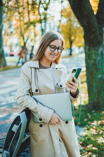 Schöne junge Frau im Herbstmantel mit Laptop und Telefon, die aus der Ferne arbeitet Frau, die am Handy im Herbstpark spricht