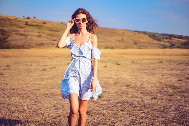 Schöne junge Frau im Kleid und in der Sonnenbrille im Freien auf dem Hügel an einem Sommertag, gegen blauen Himmel mit Wolken