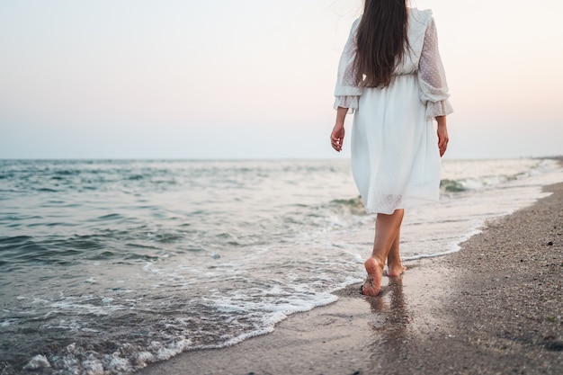 Schöne junge Frau im langen weißen Kleid mit langen Haaren und Hut geht am Strand entlang