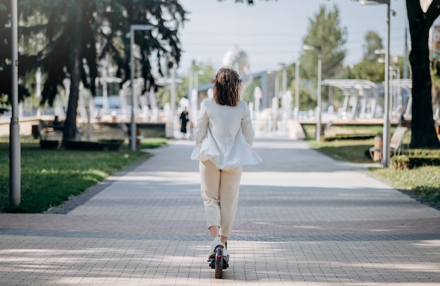 Schöne junge Frau im weißen Anzug fährt mit ihrem Elektroroller in der Stadtparklandschaft zur Arbeit