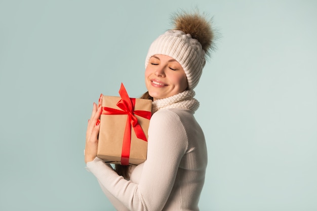 Schöne junge Frau im Winterhut mit einem Geschenk in der Hand auf einem blauen Hintergrund