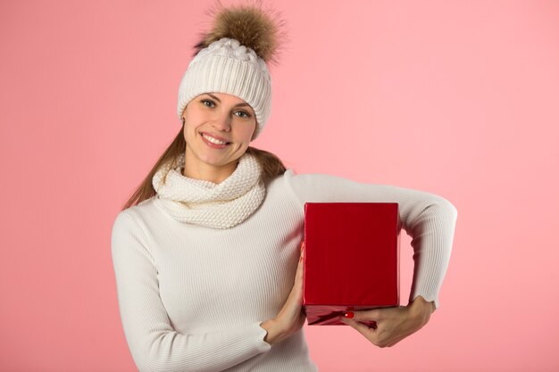 Schöne junge Frau im Winterhut mit einem Geschenk in der Hand auf einem rosa Hintergrund
