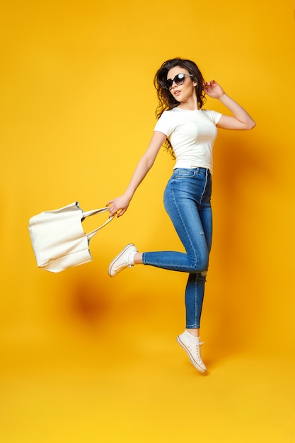 Schöne junge Frau in der Sonnenbrille, weißes Hemd, Blue Jeans, die mit Tasche auf dem gelben Hintergrund springen