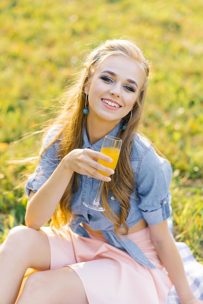 Schöne junge Frau in einem blauen Jeanshemd und rosa Rock im Garten bei einem Picknick mit einem Glas Saft in der Hand