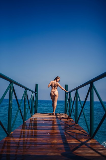 Schöne junge Frau in einem braunen Badeanzug geht an einem sonnigen Sommertag auf dem Pier am Strand am Meer