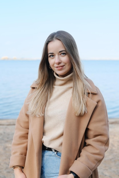 Foto schöne junge frau in einem braunen mantel posiert am strand mädchen lächelt und schaut in die kamera