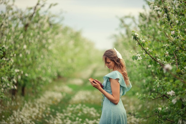 Schöne junge Frau in einem romantischen Kleid liest im Frühling in einem blühenden Garten ein zweites Buch