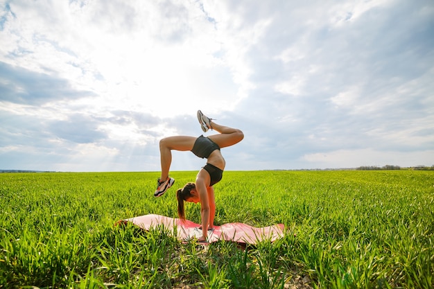 Schöne junge Frau in einem schwarzen Top und Shorts führt einen Handstand durch. Ein Model steht auf ihren Händen und macht gymnastische Spagat gegen den blauen Himmel. Gesundes Lebensstilkonzept