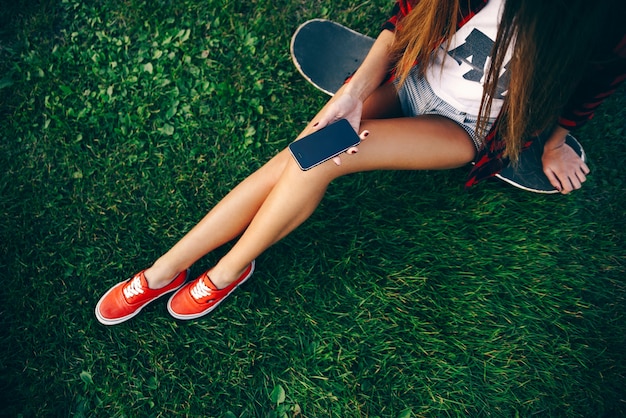 Schöne junge Frau in einem weißen T-Shirt, rotem Hemd, Shorts und Turnschuhen, die auf einem Skateboard auf Gras sitzen