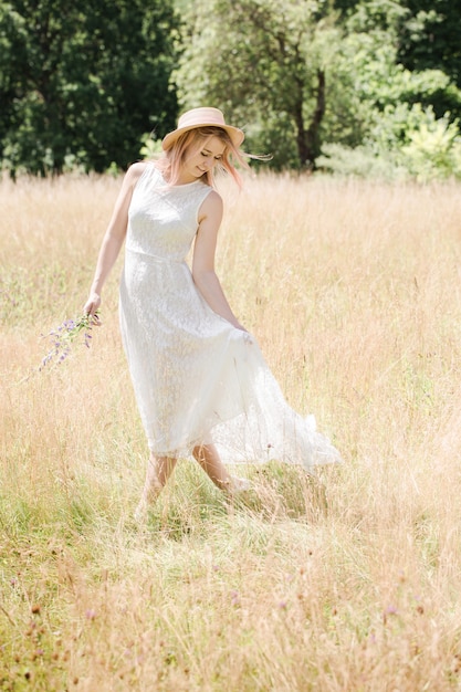 Schöne junge Frau in einem weißen Vintage-Spitzenkleid mit rosa Haaren und einem Hut geht über die Wiese